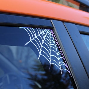 May include: A close-up of a car window with a white spiderweb decal on the corner. The car is orange and the window is tinted.