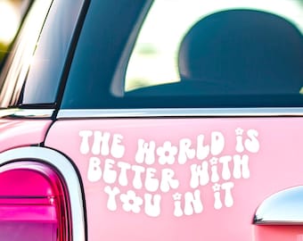 May include: A white vinyl decal applied to the rear of a pink car. The decal features the text THE WORLD IS BETTER WITH YOU IN IT in a rounded, retro-style font with small flower icons integrated into the lettering, positioned near the taillight.