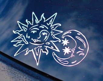 May include: A white vinyl decal applied to a dark car window, featuring a stylised sun with a human face and a crescent moon with a human face, accompanied by two small four-pointed stars positioned between the celestial bodies.