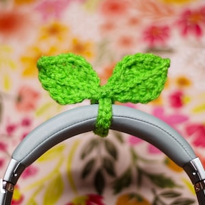 May include: Green crocheted leaf-shaped headphone covers, labeled "SIZE: SMALL", on a pair of gray headphones against a floral background.