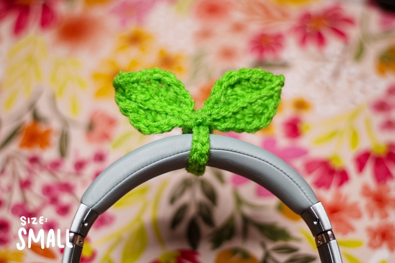 May include: Green crocheted leaf-shaped headphone covers, labeled "SIZE: SMALL", on a pair of gray headphones against a floral background.