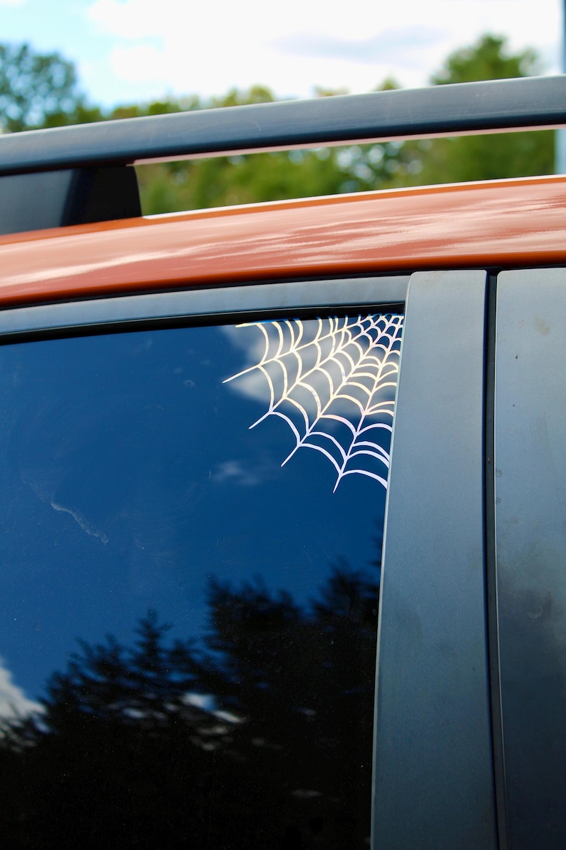 May include: A white spider web decal applied to the upper corner of a dark tinted vehicle window. The graphic features a series of radiating lines connected by curved segments, creating a classic web pattern that contrasts against the glass.