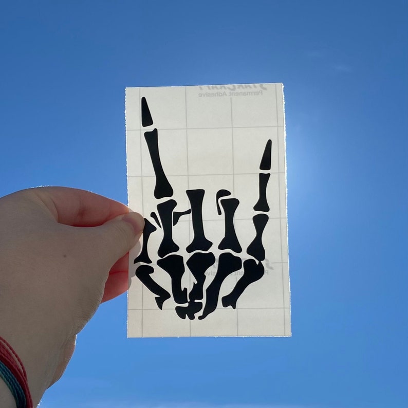 K&ouml;nnte beinhalten: Eine schwarze Skeletthand, die die Rock'n'Roll-Handgeste macht. Die Hand befindet sich auf einem transparenten Blatt Transferpapier.