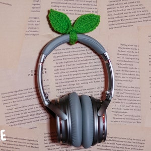 May include: A pair of gray noise-canceling headphones with a brown headband. The headphones are sitting on a stack of books with a green crocheted leaf design on top.