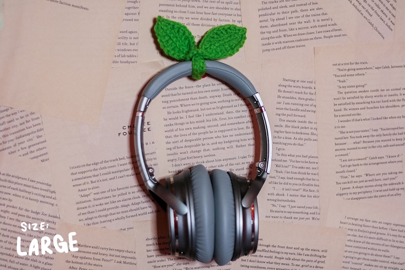 May include: A pair of gray noise-canceling headphones with a brown headband. The headphones are sitting on a stack of books with a green crocheted leaf design on top.