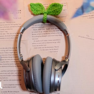 May include: Gray over-ear headphones with a crocheted green plant attached to the top. The headphones are lying on a stack of books with text.