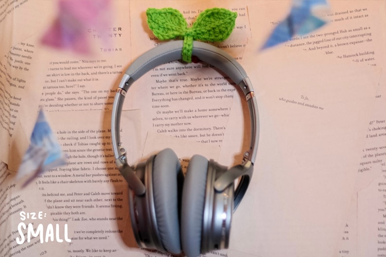May include: Gray over-ear headphones with a crocheted green plant attached to the top. The headphones are lying on a stack of books with text.
