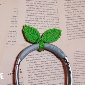 May include: A pair of gray headphones with a green crocheted leaf design. The headphones are resting on a book with text. The text on the book reads "SIZE: LARGE".