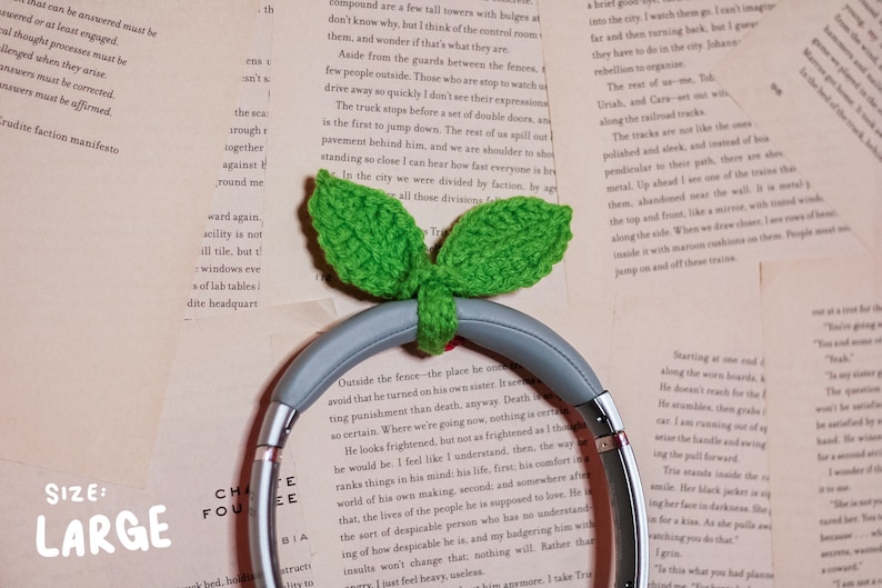 May include: A pair of gray headphones with a green crocheted leaf design. The headphones are resting on a book with text. The text on the book reads "SIZE: LARGE".