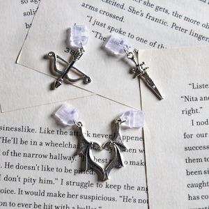May include: Three silver earrings with different charms: a bow and arrow, a sword, and a heart with wings. The earrings are hanging from clear plastic earring backs.
