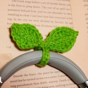 May include: A small, green crocheted leaf design headphone holder. The holder is attached to a pair of gray headphones. The text "SIZE: SMALL" is visible in the lower left corner of the image.