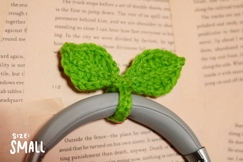 May include: A small, green crocheted leaf design headphone holder. The holder is attached to a pair of gray headphones. The text "SIZE: SMALL" is visible in the lower left corner of the image.