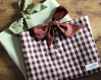 May include: Two gingham placemats with bow accents. One placemat is green and white, the other is brown and white. The brown and white placemat has a small white label that says "Sunday in Maine". The bows are made of a solid color fabric.