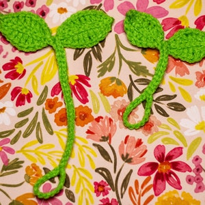 May include: Two green crocheted leaf bookmarks with a stem. The bookmarks are on a pink background with a floral pattern.