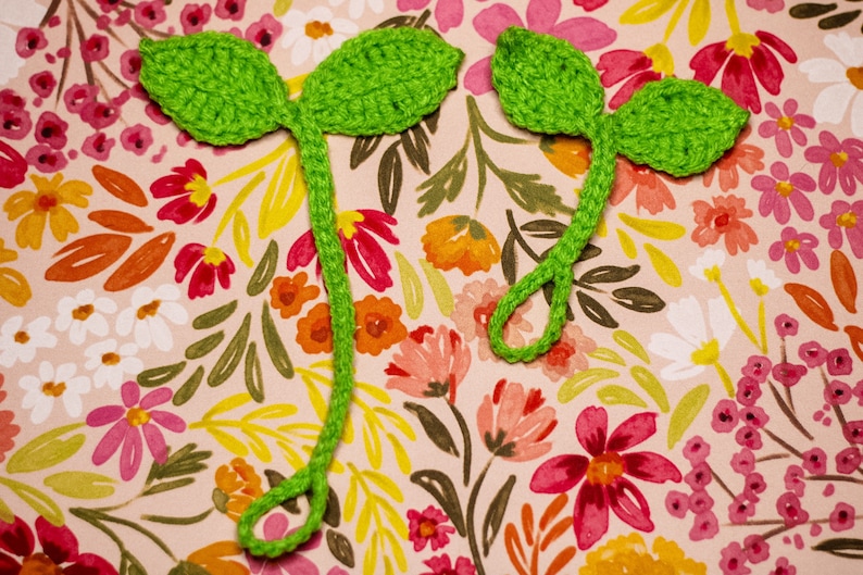 May include: Two green crocheted leaf bookmarks with a stem. The bookmarks are on a pink background with a floral pattern.