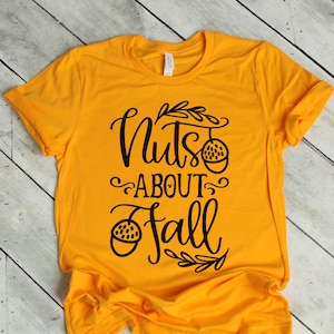 May include: A yellow t-shirt with the text "Nuts About Fall" in black lettering. The text is in a handwritten font and includes a small acorn graphic.