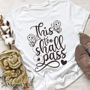 May include: White t-shirt with a black graphic that says "This too shall pass" with two butterflies and hearts.