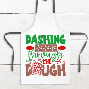 May include: White apron with grey straps and the text "Dashing through the dough" in green, red, and brown. A rolling pin with snowflakes is above the text.