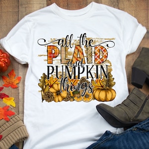May include: White t-shirt with the text "all the plaid and pumpkin things" in black lettering. The text is surrounded by a gold glitter background and a fall-themed design with pumpkins, leaves, and pinecones.