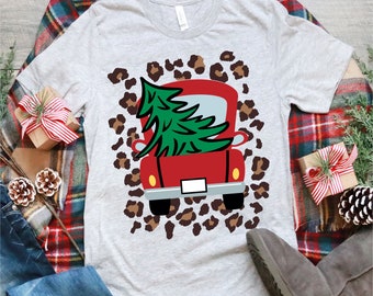 Könnte beinhalten: Ein roter Pickup mit einem grünen Weihnachtsbaum auf der Ladefläche, auf einem weißen T-Shirt mit Leopardenmuster.