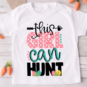 Peut inclure: T-shirt blanc avec le texte "this girl can hunt" en noir, rose et turquoise. Le mot "girl" est décoré de pois roses et blancs. Le texte est accompagné d'une flèche et d'un cœur.