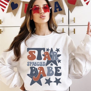 May include: White sweatshirt with the text "STAR SPANGLED BABE" in retro font and star graphics in red, blue, and brown. The sweatshirt is a casual style, perfect for patriotic events.