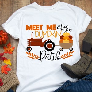 May include: White t-shirt featuring a brown tractor pulling a wagon full of orange pumpkins. The text "Meet me at the pumpkin patch" is printed on the shirt.