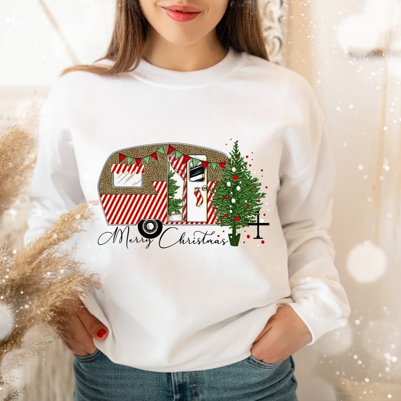 May include: White sweatshirt with a red and white striped camper with a Christmas tree next to it. The camper has a brown and gold leopard print roof and a green and red garland. The text "Merry Christmas" is printed below the camper.