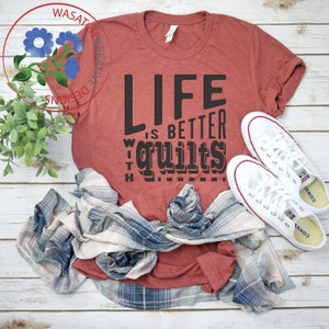 May include: A red t-shirt with the text "Life is better with quilts" printed on it. The shirt is laid out on a wooden surface with a plaid shirt and white sneakers.