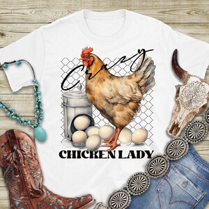 May include: White t-shirt featuring a brown chicken illustration and the text "Crazy Chicken Lady". The chicken is standing on a wire fence with a basket of eggs beside it.