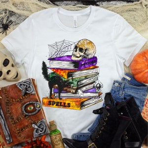 以下が含まれることがあります： 白いTシャツに、黒い猫、頭蓋骨、そして「Spells」と「Potions」と書かれた本が積み重ねられています。本は蜘蛛の巣で囲まれています。