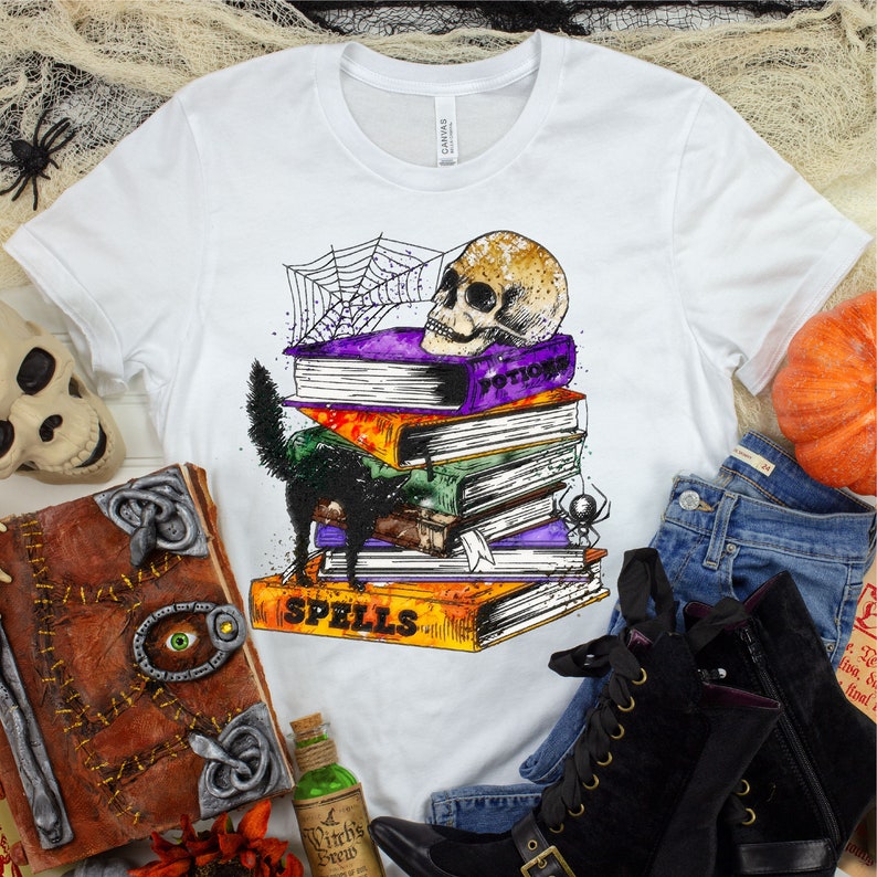 以下が含まれることがあります： 白いTシャツに、黒い猫、頭蓋骨、そして「Spells」と「Potions」と書かれた本が積み重ねられています。本は蜘蛛の巣で囲まれています。