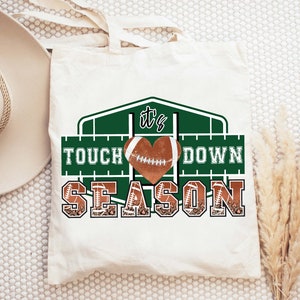 Peut inclure: Un sac fourre-tout blanc avec un motif graphique vert et marron. Le graphique repr&eacute;sente un ballon de football en forme de c&oelig;ur avec le texte "It's Touchdown Season".