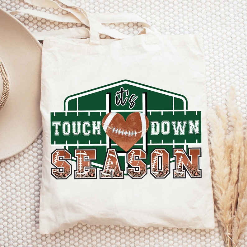 Peut inclure: Un sac fourre-tout blanc avec un motif graphique vert et marron. Le graphique repr&eacute;sente un ballon de football en forme de c&oelig;ur avec le texte "It's Touchdown Season".