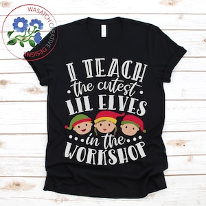 May include: Black t-shirt with white text: "I teach the cutest lil elves in the workshop." The design features three cartoon elf faces wearing red, green, and yellow hats. The Wasatch Creative Designs logo is in the upper left corner.
