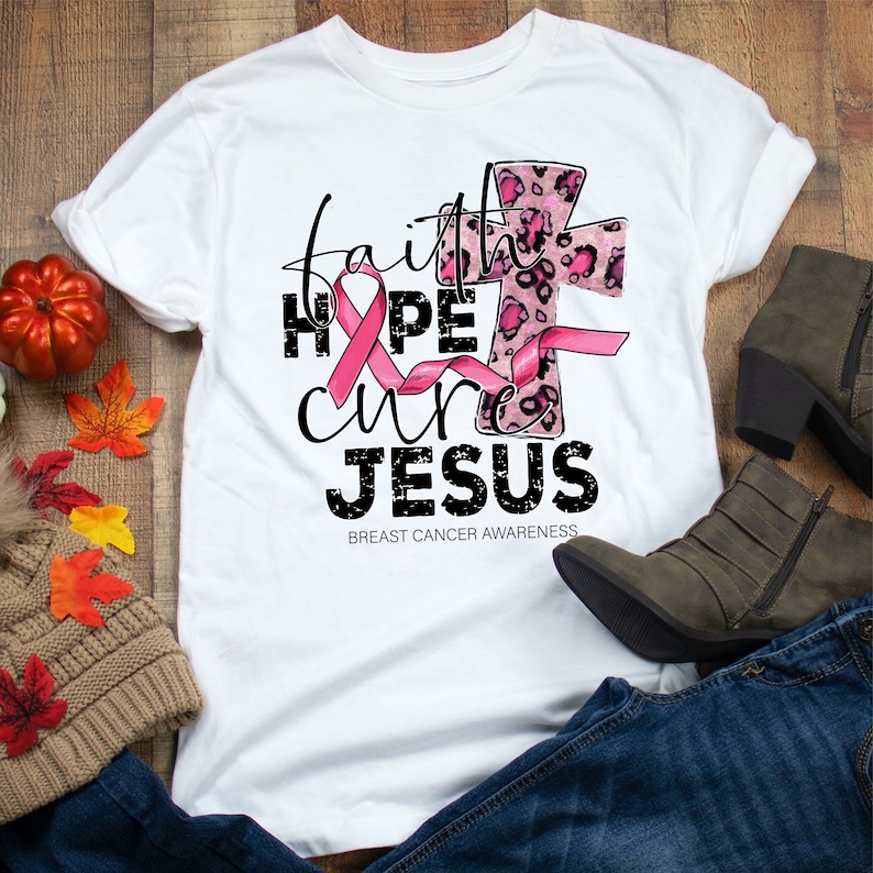 May include: White t-shirt with a pink ribbon, a leopard print cross, and the text "faith hope cure JESUS" in black with a distressed effect. The text "BREAST CANCER AWARENESS" is printed below the main text.