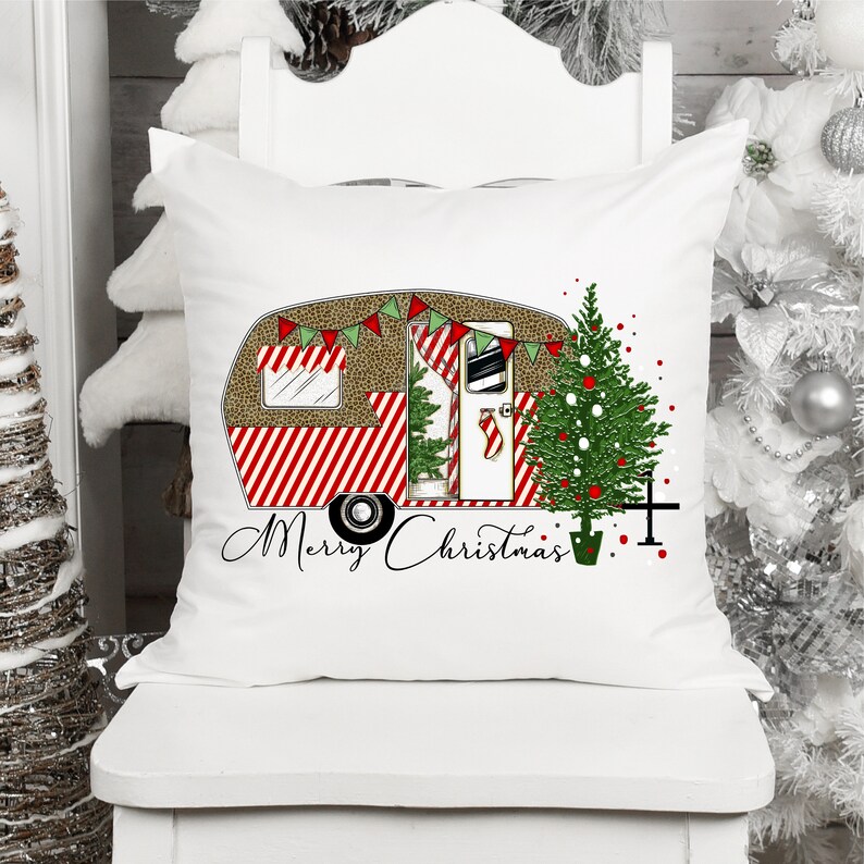 May include: White throw pillow with a red and white striped camper with a Christmas tree next to it. The camper has a brown and white leopard print roof and a green garland with red and white flags. The text "Merry Christmas" is written below the camper.