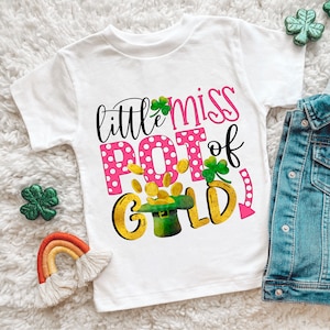 May include: White t-shirt with a pink and white polka dot design and the text "little miss pot of gold" in gold and green lettering. The design includes a leprechaun's hat with gold coins.