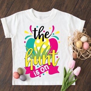 Peut inclure: T-shirt blanc avec un motif graphique coloré représentant des œufs, de l'herbe et le texte "the hunt is on".