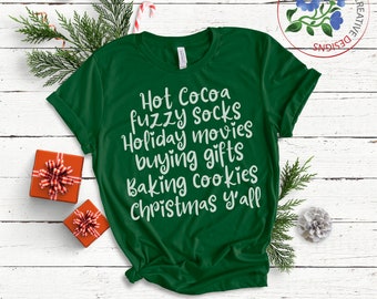 Könnte beinhalten: Ein grünes T-Shirt mit weißem Text, der "Hot Cocoa fuzzy socks Holiday movies buying gifts Baking cookies Christmas y'all" lautet.