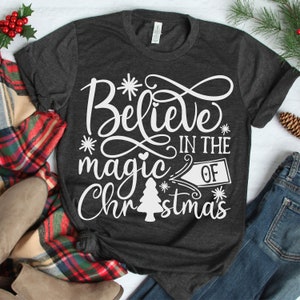 May include: A dark gray t-shirt with a white design that says "Believe in the magic of Christmas". The design features a stylized Christmas tree and snowflakes.