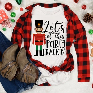 May include: White t-shirt featuring a red and green nutcracker illustration and the text "Let's get this party crackin'" on it. The shirt is layered over a red and black checked shirt.