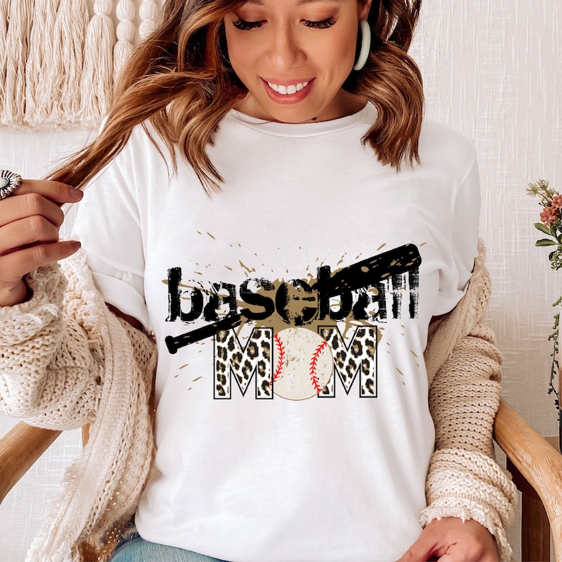 Peut inclure: T-shirt blanc avec un motif graphique noir et marron qui dit "baseball mom" avec un ballon de baseball et une batte de baseball.