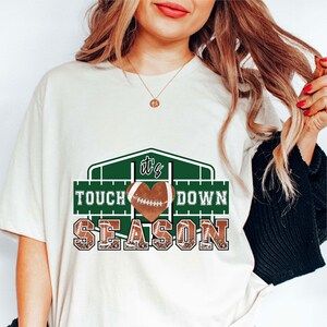 Peut inclure: T-shirt blanc avec un motif graphique vert et marron. Le graphique repr&eacute;sente un ballon de football en forme de c&oelig;ur avec le texte "It's Touch Down Season".