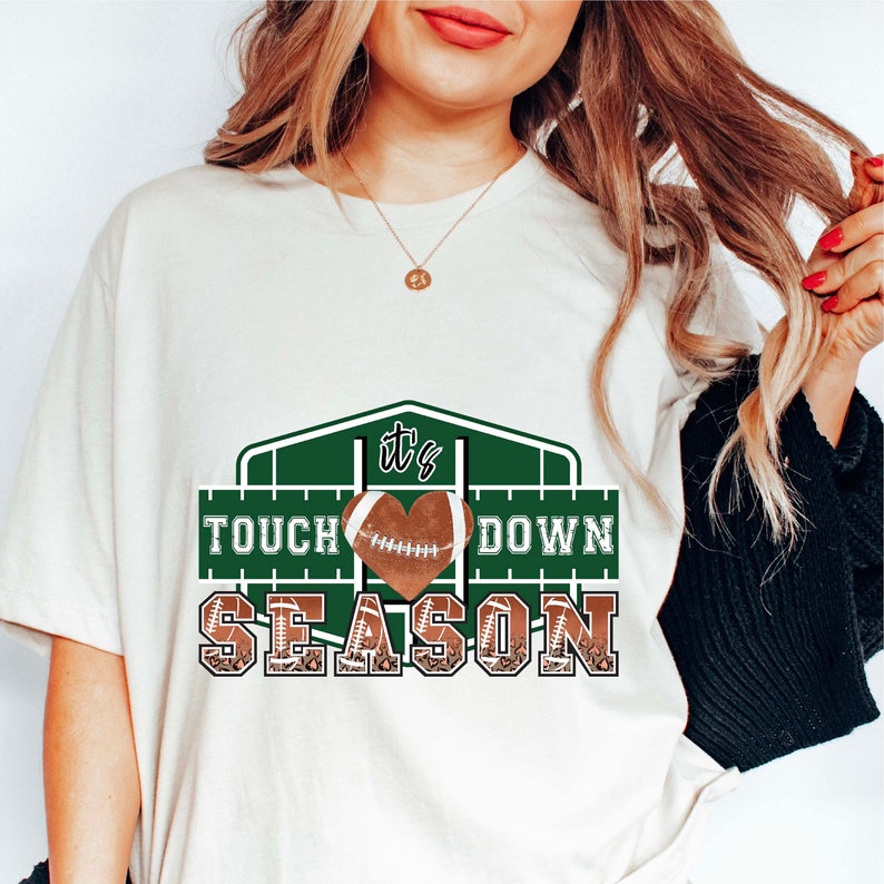 Peut inclure: T-shirt blanc avec un motif graphique vert et marron. Le graphique repr&eacute;sente un ballon de football en forme de c&oelig;ur avec le texte "It's Touch Down Season".
