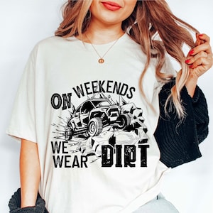 May include: White t-shirt with a black graphic design of an off-road vehicle driving through rocks. The text "On Weekends We Wear Dirt" is printed on the shirt.