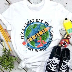 Puede incluir: Camiseta blanca con un globo terráqueo acuarela azul y verde con un lápiz amarillo que lo atraviesa. El texto alrededor del globo dice "Es un gran día para enseñar a los pequeños cambiadores del mundo".