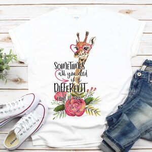 May include: White t-shirt with a watercolor illustration of a giraffe wearing pink heart-shaped glasses and a floral arrangement. The text "Sometimes all you need is a DIFFERENT point of view" is printed on the shirt.
