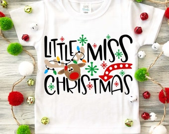 Puede incluir: Un body blanco para bebé con un gráfico de reno negro y rojo y el texto "Little Miss Christmas" en negro. El reno tiene cuernos rojos y una nariz roja. El texto está rodeado de copos de nieve verdes y blancos.