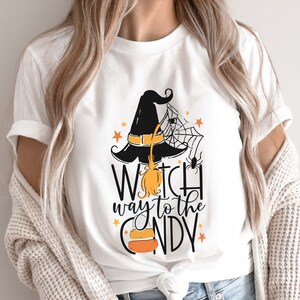 May include: White t-shirt with a black witch hat, broom, spider web, and stars. The text "Witch way to the candy" is printed on the shirt.